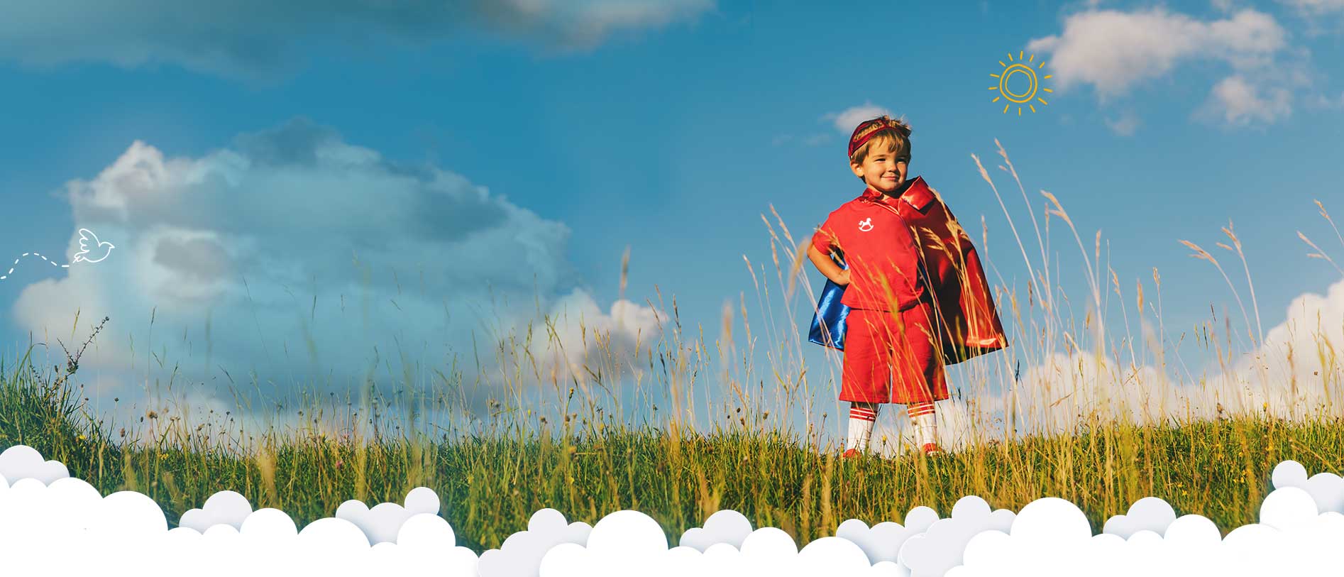 A boy standing in a field wearing a red tshirt, shorts and cape in a super hero pose